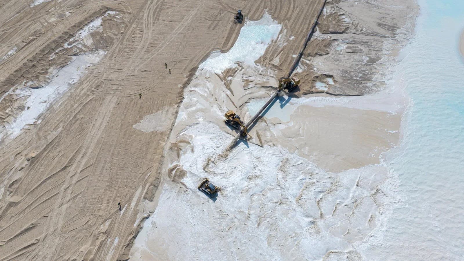 Aerial view of construction machinery spreading sand on reclaimed land beside pale turquoise water